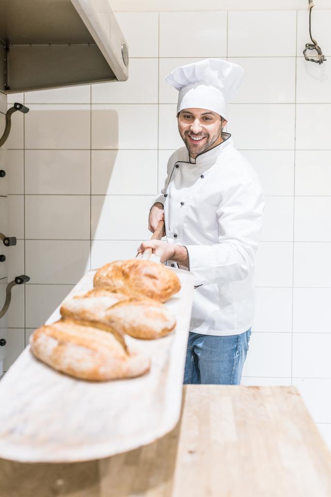 Panadero sosteniendo pala de metal con pastelería fresca en la cocina panadería
 - Foto, Imagen