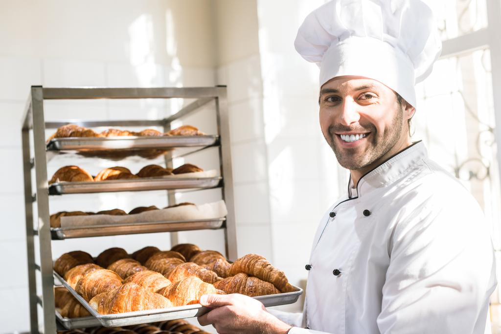 Smiling baker holding tray with fresh cooked croissants in bakehouse - Photo, Image
