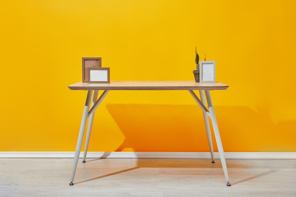 Table en bois avec cadres photo et cactus près du mur jaune
 - Photo, image