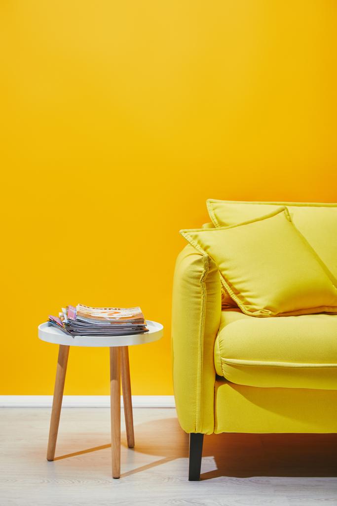 Καναπές με μαξιλάρια και μικρό τραπέζι με περιοδικά κοντά κίτρινο τοίχο - Φωτογραφία, εικόνα