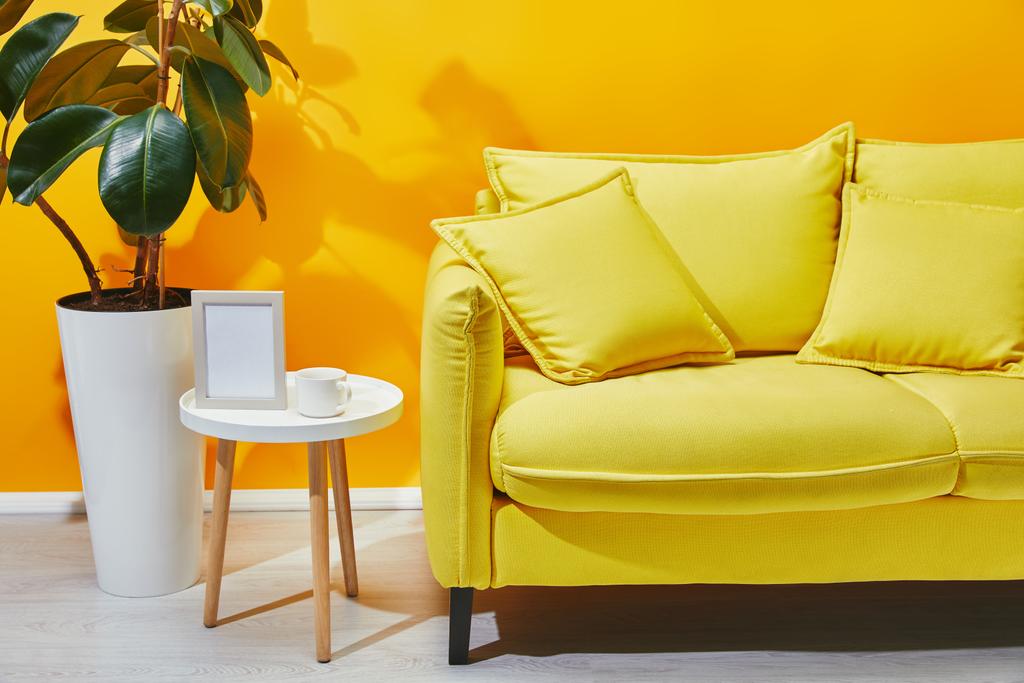 Sofá, ficus e pequena mesa com moldura de foto e xícara de café perto da parede amarela
 - Foto, Imagem