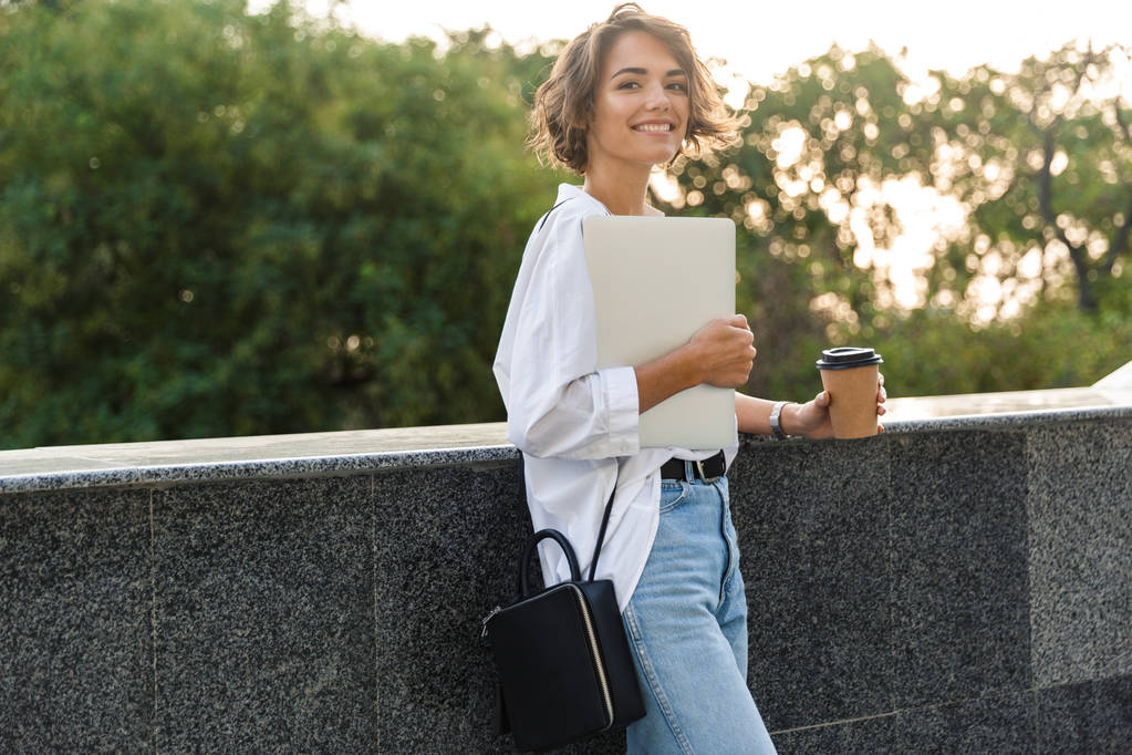 Foto von jungen netten schönen Frau zu Fuß im Freien mit Laptop-Computer hält Kaffee. - Foto, Bild