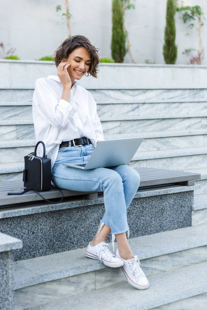 Foto van jonge mooie vrouw zitten buiten met behulp van laptop computer luisteren muziek met koptelefoon. - Foto, afbeelding