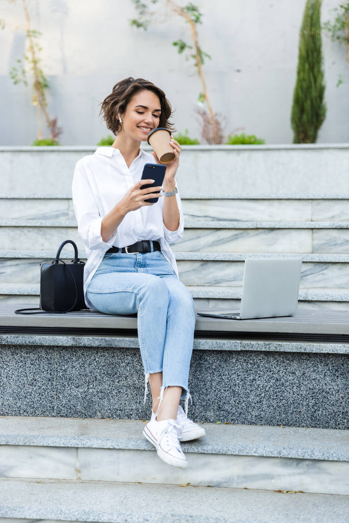 Sorrindo jovem mulher em fones de ouvido sentado ao ar livre na rua, falando no celular, bebendo café
 - Foto, Imagem