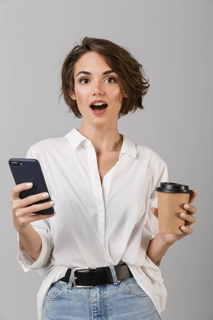 Foto van geschokt jonge zakenvrouw poseren geïsoleerd over grijs muur achtergrond chatten via de mobiele telefoon koffie drinken - Foto, afbeelding