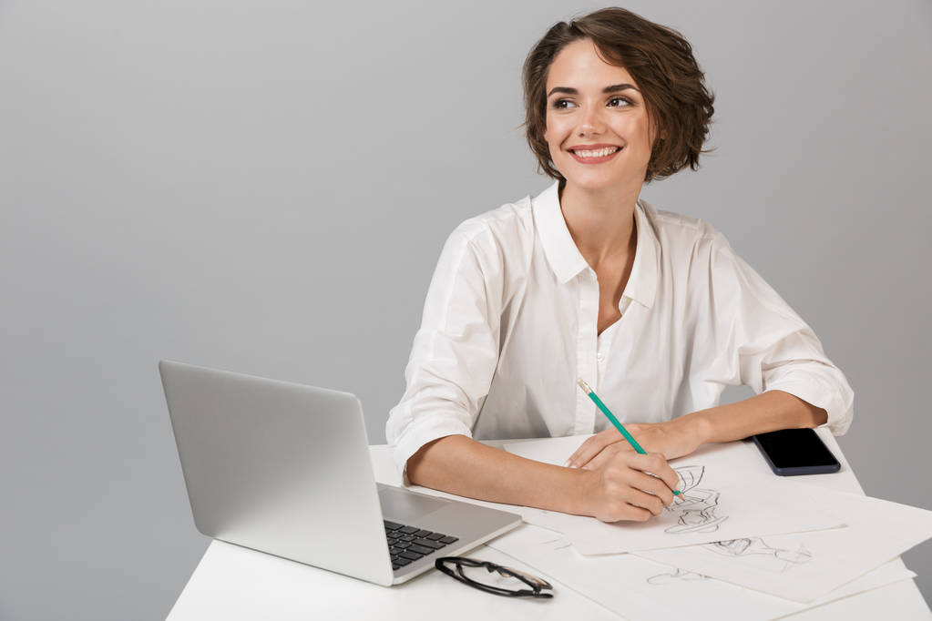 Beeld van jonge zakenvrouw poseren geïsoleerd over grijs muur achtergrond zitten aan de tafel met behulp van laptop schrijven van notities. - Foto, afbeelding