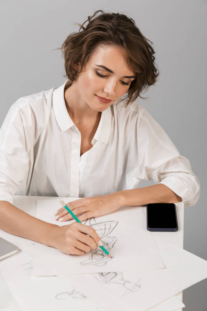 Зображення молодої ділової жінки, яка позує ізольовано на сірому фоні стіни, сидить за столом, використовуючи малюнок ноутбуків
. - Фото, зображення