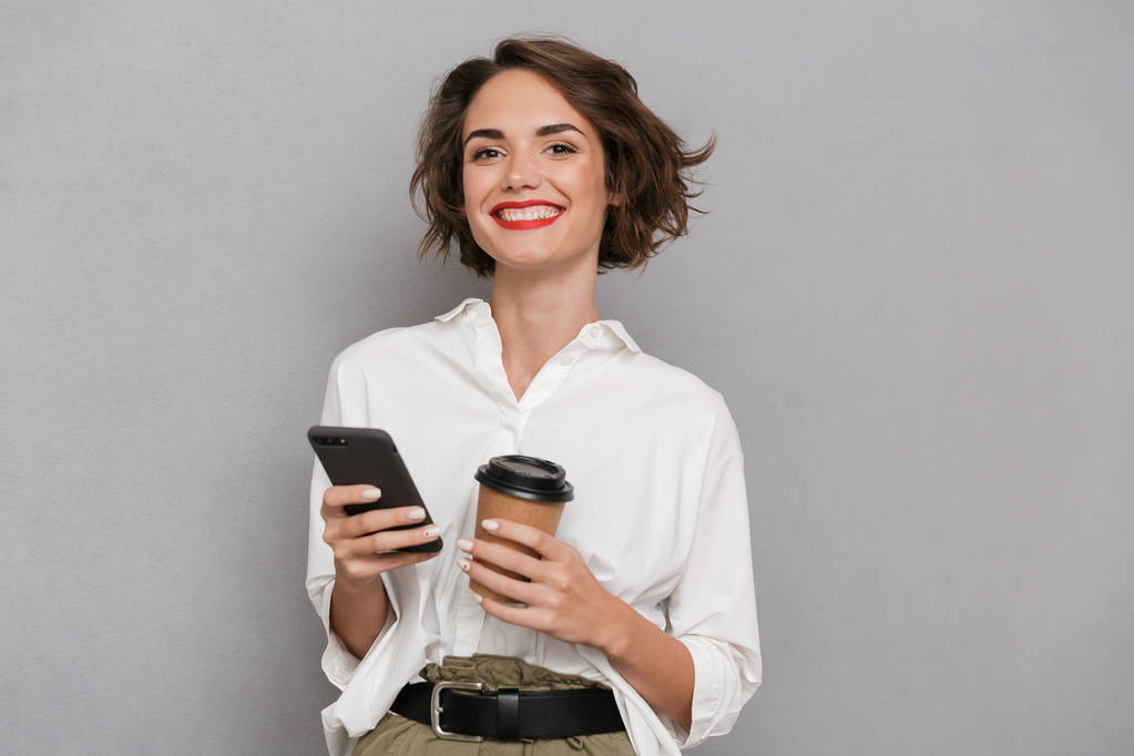 Foto da mulher satisfeita 20s segurando takeaway café e usando telefone celular isolado sobre fundo cinza
 - Foto, Imagem