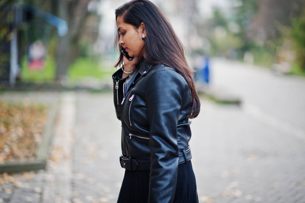 Досить індійська дівчина в чорній сукні сарі і шкіряній куртці позують на вулиці осінь і розмовляють на мобільному телефоні
. - Фото, зображення