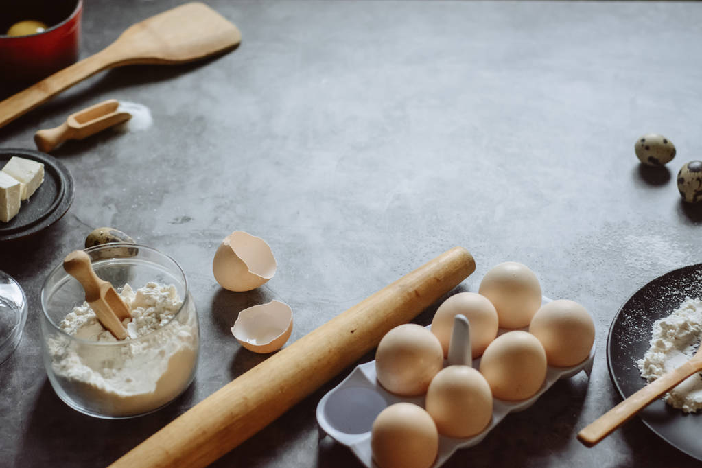 Zutaten und Utensilien zum Backen. Löffel mit Mehl, Geschirr, Eiern, Buttersalz und Nudelholz auf grauem Hintergrund. flach lag. Textraum - Foto, Bild