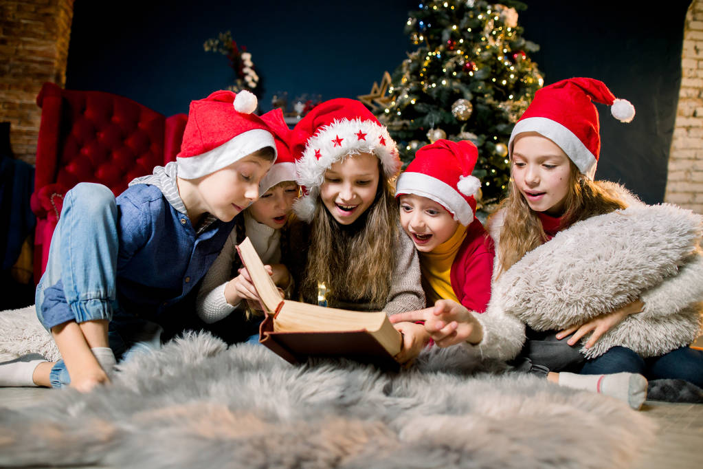 Charmante en prachtige lieve kinderen in Santa's hoeden kerst verhalen lezen en lachen, op de achtergrond van een kerstboom. - Foto, afbeelding