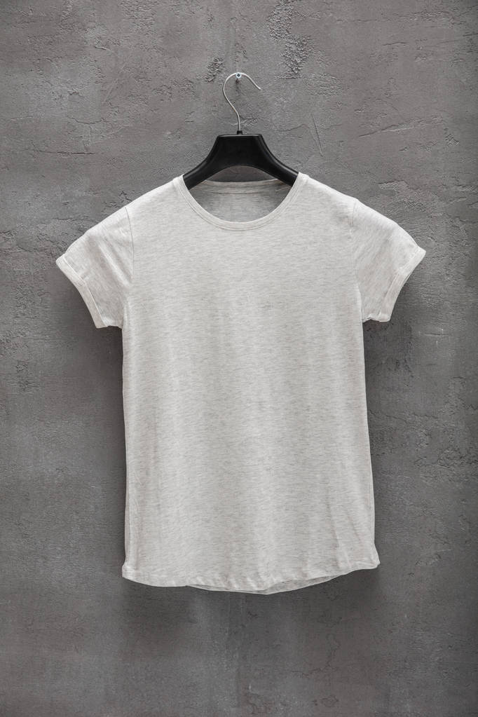 Voorzijde van vrouwelijke grijs melange katoen t-shirt op een hanger en een betonnen wand in de achtergrond. T-shirt zonder afdrukken - Foto, afbeelding