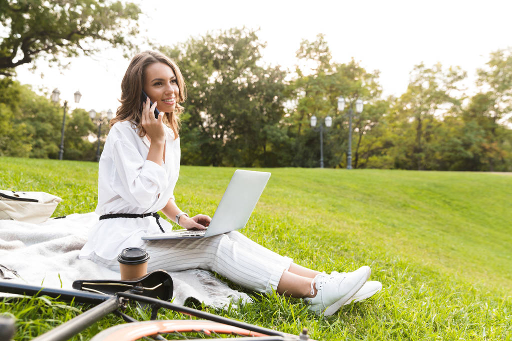Mooie jonge vrouw fietser tijd doorbrengen in het park, zittend op een gras, met behulp van laptopcomputer, praten op mobiele telefoon - Foto, afbeelding