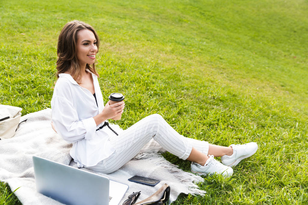 S úsměvem mladá žena tráví čas v parku, sedí na trávě s notebookem, držící šálek kávy - Fotografie, Obrázek
