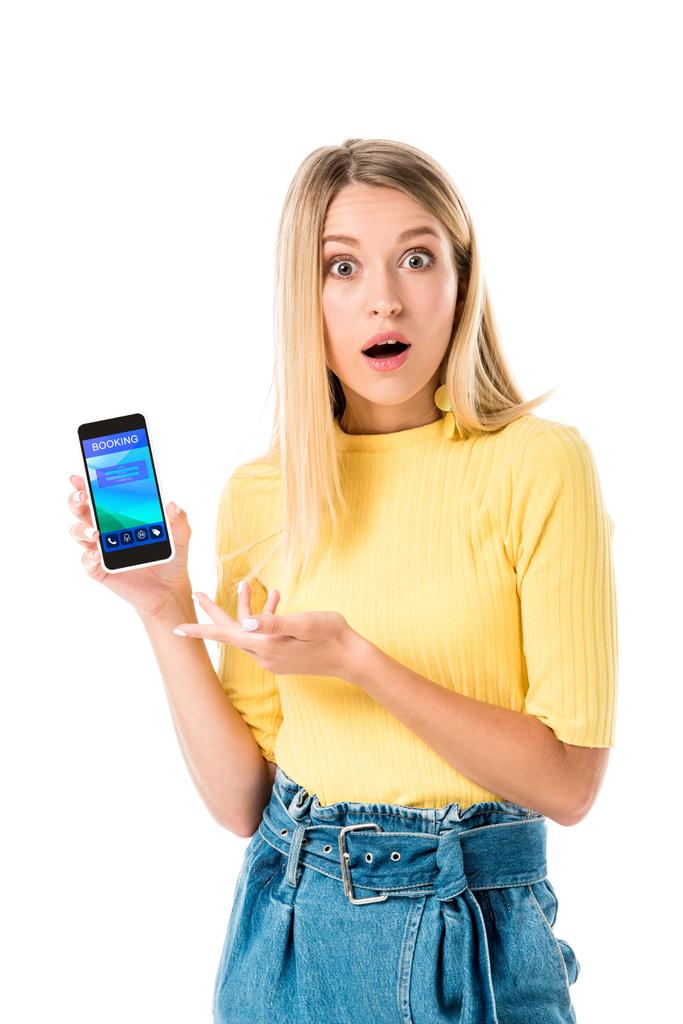 шокированная молодая женщина, держащая смартфон с приложением бронирования на экране и глядя на камеру, изолированную на белом
  - Фото, изображение