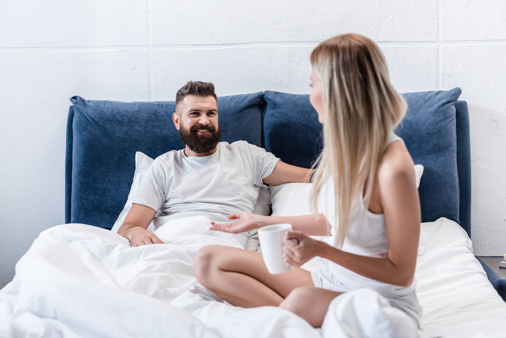 chica rubia con taza de café hablando con el hombre barbudo sonriente acostado en almohadas blancas en la cama
 - Foto, Imagen