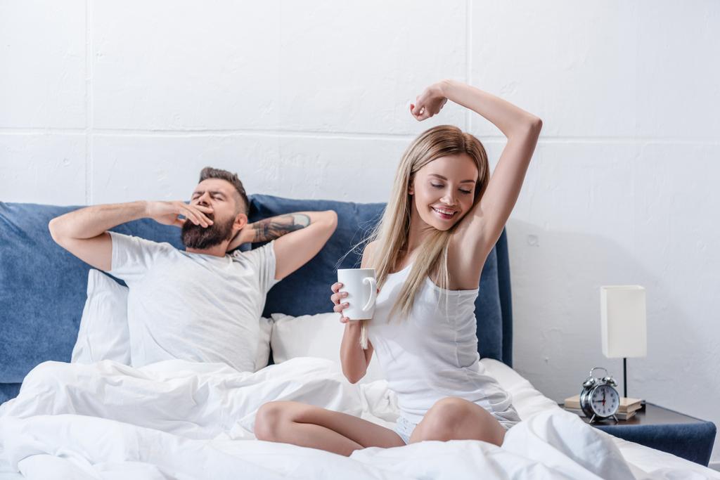 bärtiger Mann gähnt, während attraktive junge Frau mit Kaffeetasse sich im Bett streckt - Foto, Bild