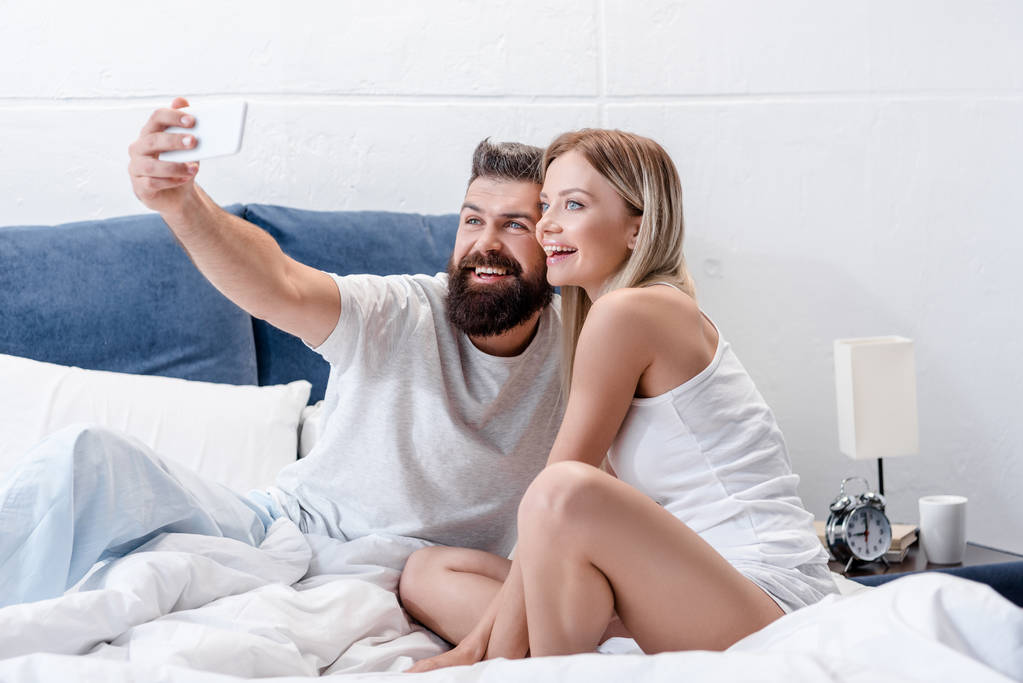 Бородатый мужчина и красивая девушка улыбаются и делают селфи в белой постели утром
 - Фото, изображение