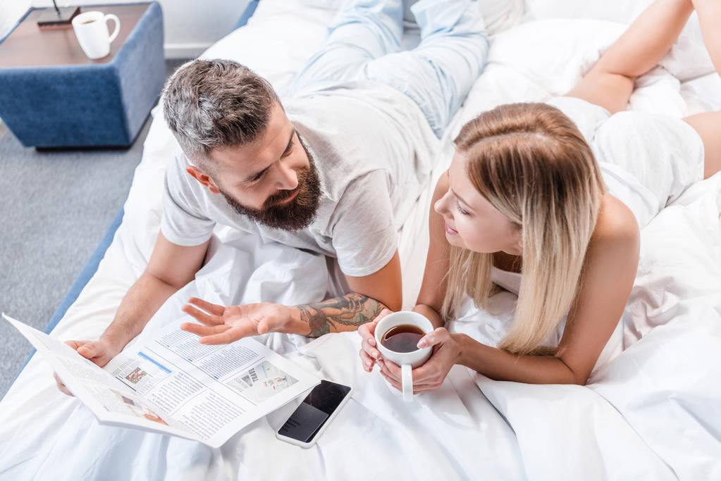 bärtiger Mann mit Zeitung und attraktive Frau mit Kaffeetasse liegen im Bett und reden - Foto, Bild