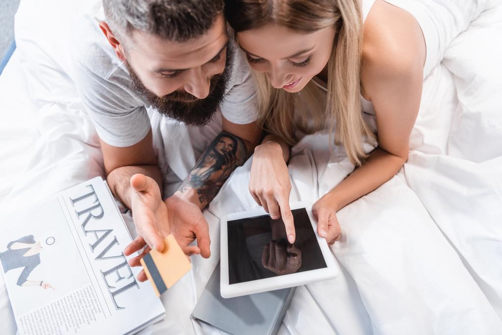 Casal jovem com cartão de crédito e tablet digital deitado em edredão branco e fazendo compras on-line
 - Foto, Imagem