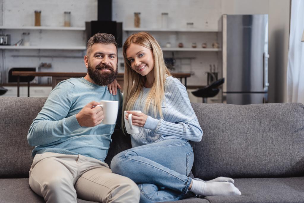 glückliches Paar sitzt in lässiger Kleidung mit Getränken auf dem Sofa - Foto, Bild