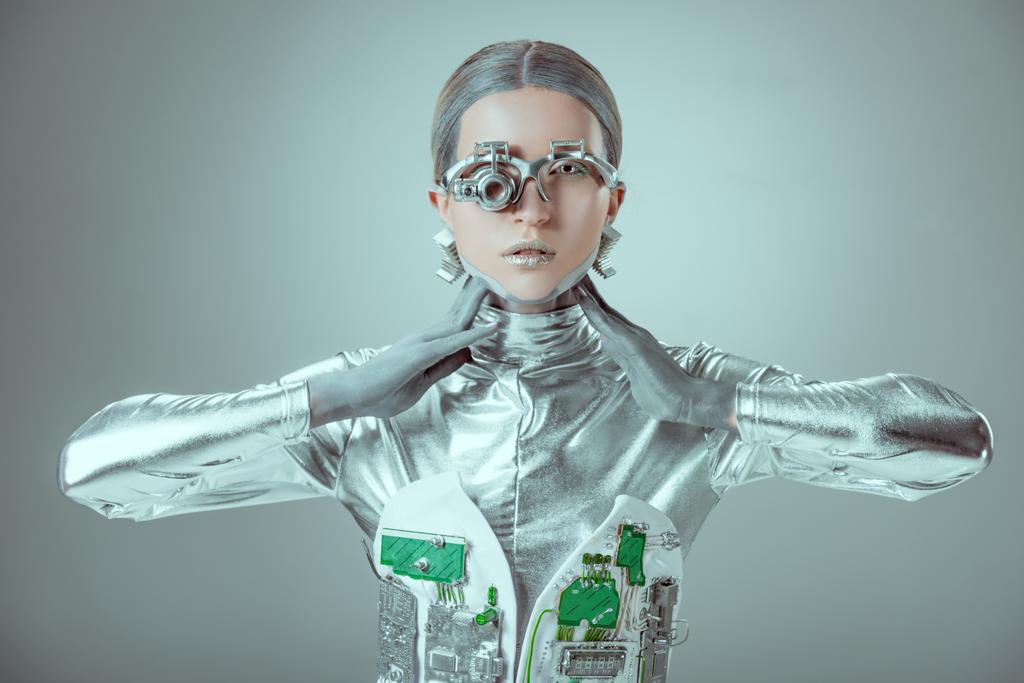 junge Roboterin berührt Hals und blickt auf Kamera isoliert auf graues, zukünftiges Technologiekonzept  - Foto, Bild