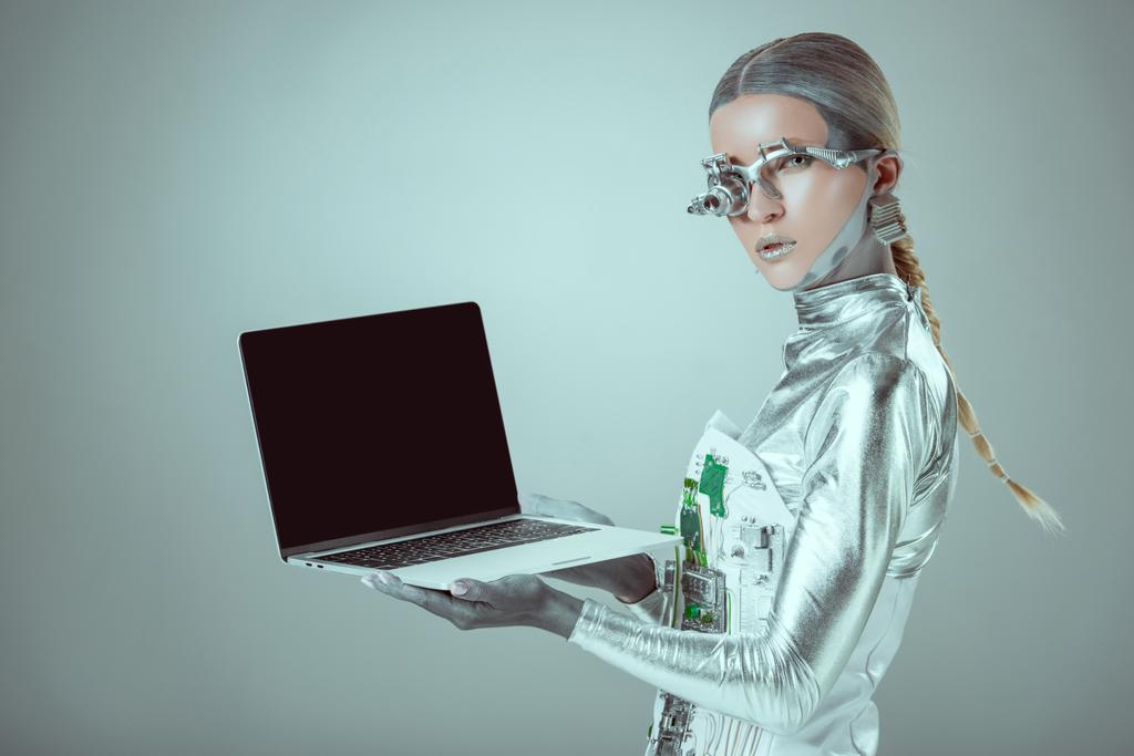 silberner Roboter hält Laptop mit leerem Bildschirm isoliert auf grauem, zukünftigem Technologiekonzept - Foto, Bild