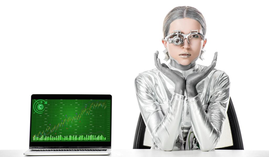 Zilveren robot zittend aan tafel in de buurt van laptop met grafiek toestel geïsoleerd op wit, toekomstige technologie concept - Foto, afbeelding