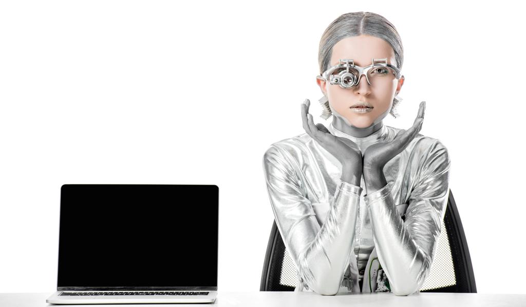 robô de prata sentado à mesa perto de laptop com tela em branco isolado em branco, conceito de tecnologia futuro
 - Foto, Imagem