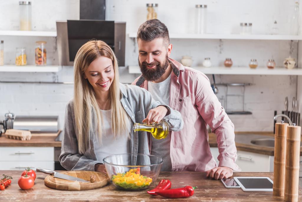 Beautiful wife standing with husband in kitchen and adding oil in salad - Photo, Image