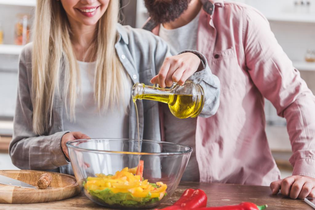 обрезанный вид улыбающейся женщины, стоящей рядом с мужем и добавляющей масло в салат в миске
 - Фото, изображение