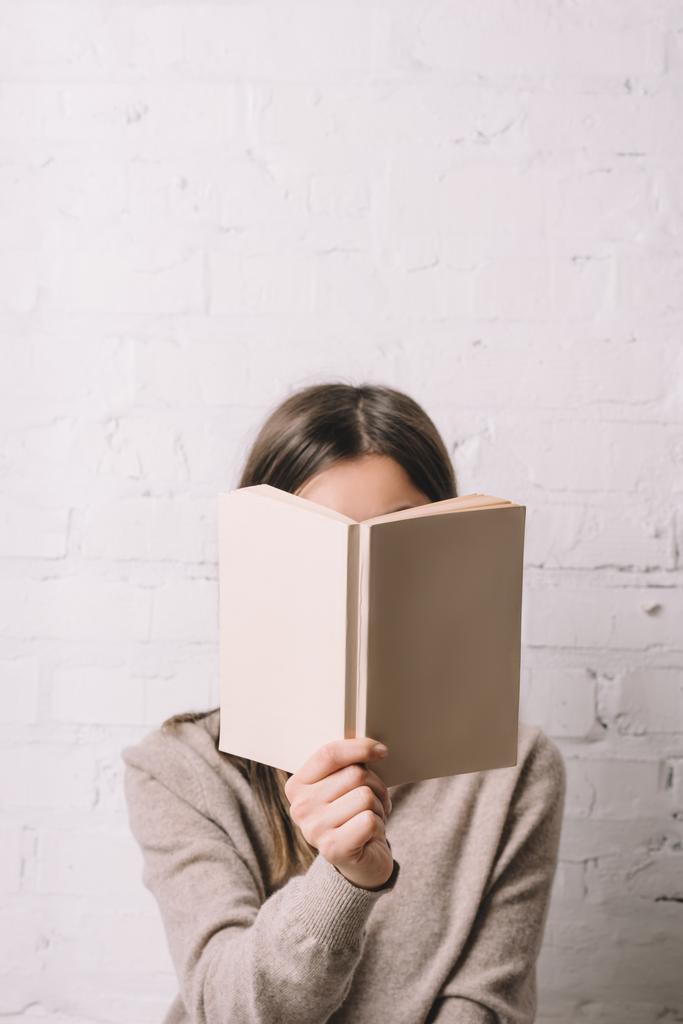 girl hiding face behind book near white brick wall - Photo, Image