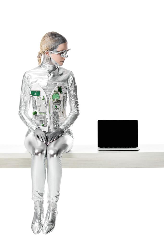 Silver robot sitting on table near laptop with blank screen isolated on white, future technology concept
 - Фото, изображение