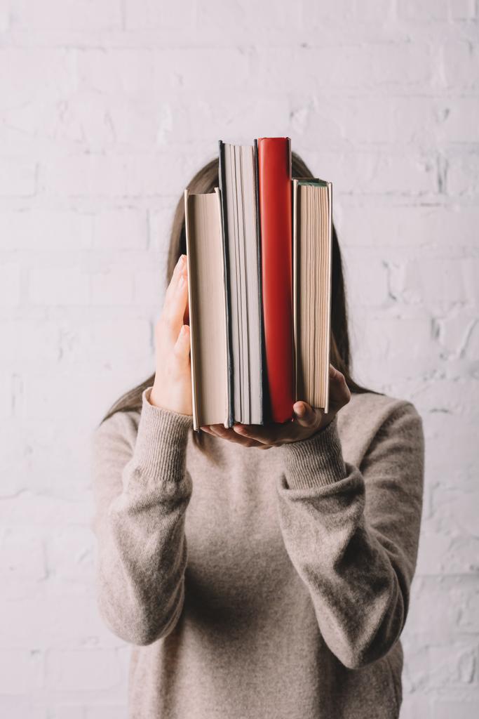 junge Frau versteckt Gesicht hinter Büchern nahe weißer Backsteinmauer - Foto, Bild
