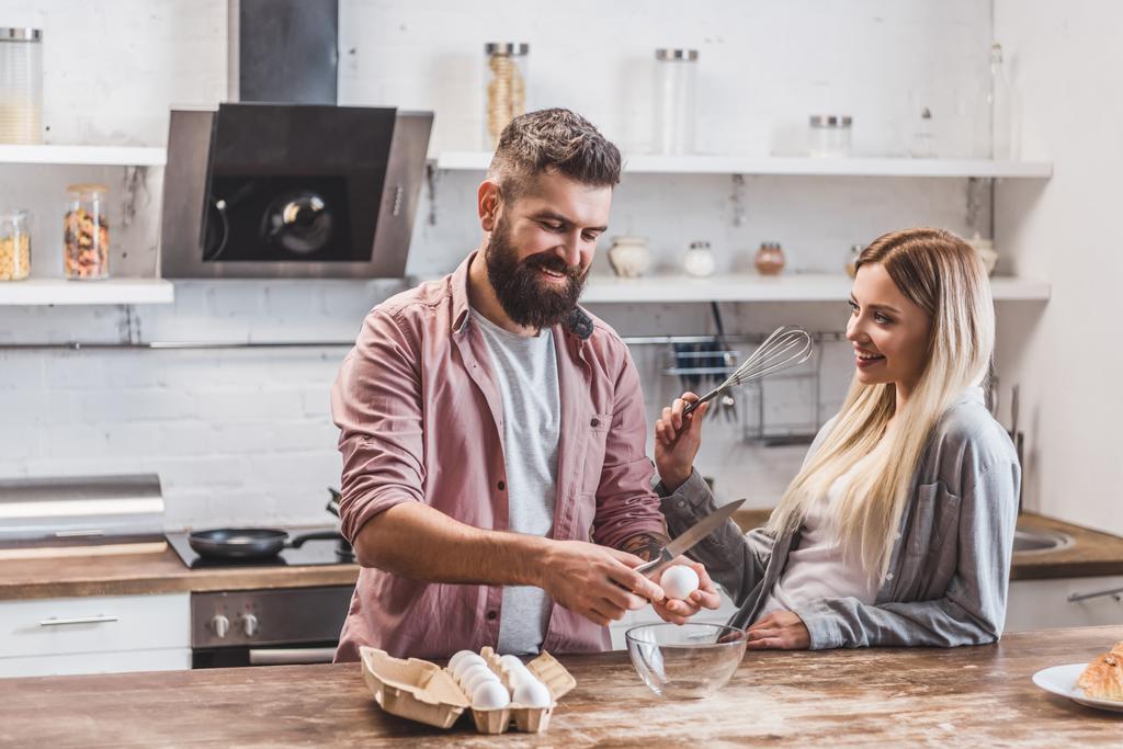 handsome bearded man smashing eggs at wooden table while woman holding balloon whisk - Photo, Image