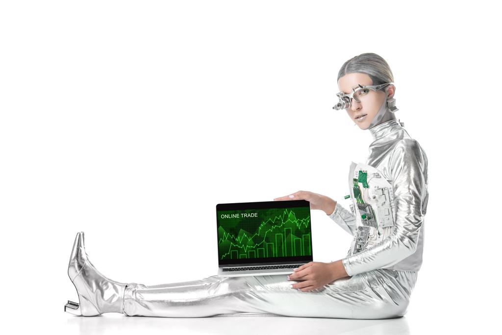 robot plateado sentado y mostrando portátil con aparato de comercio en línea aislado en blanco, concepto de tecnología futura
 - Foto, Imagen