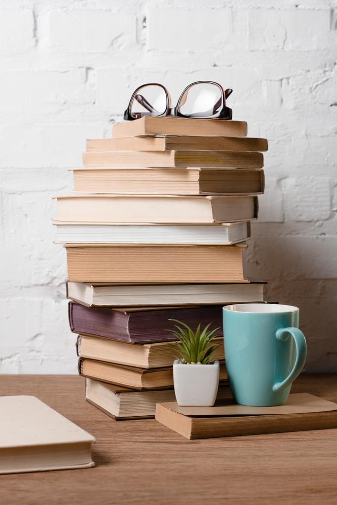 груда книг, очков, горшок с растением и чашка с горячим напитком на деревянном столе
 - Фото, изображение