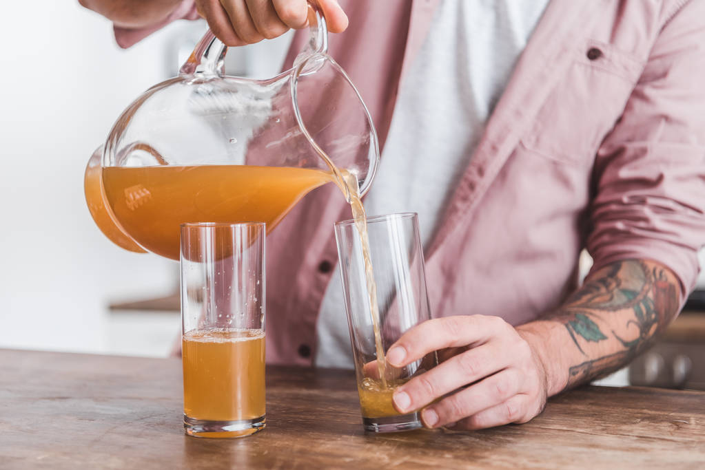 обрезанный вид человека, наливающего сок из банки в стаканы за деревянным столом
 - Фото, изображение