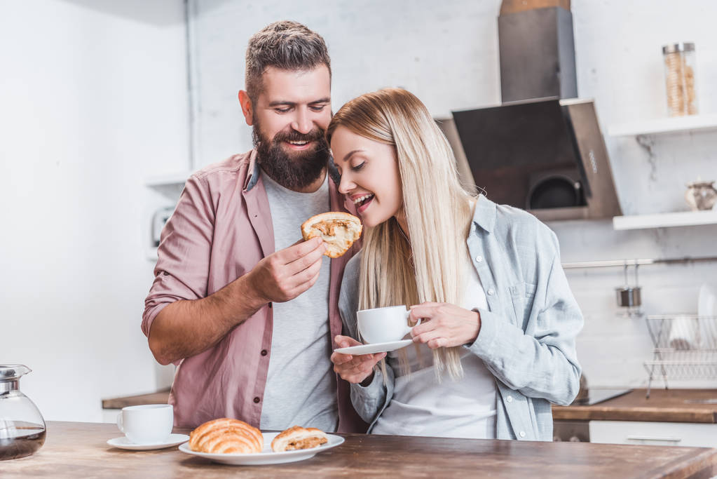 Bärtiger Mann füttert Freundin in Küche mit Croissant - Foto, Bild