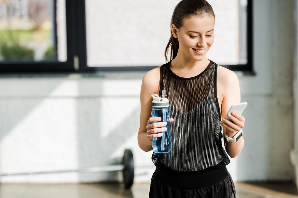 deportista sonriente sosteniendo la botella de deporte con agua y utilizando el teléfono inteligente en el gimnasio
 - Foto, Imagen