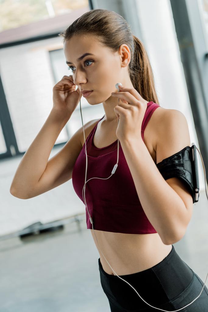 concentrated sportswoman in smartphone armband putting on earphones at gym - Photo, Image