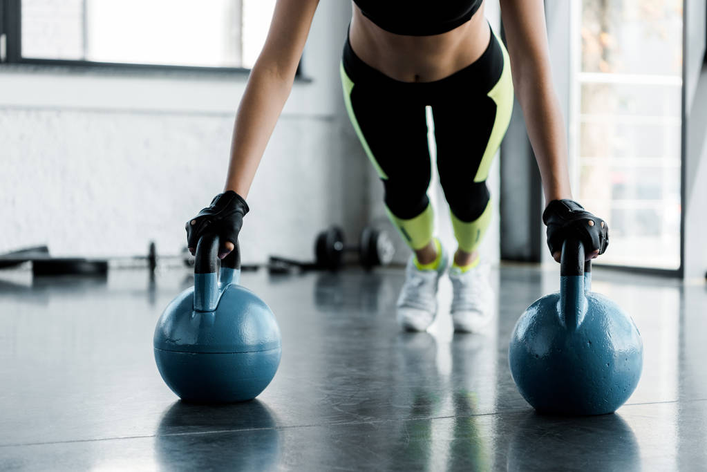 Ausgeschnittene Ansicht einer fitten Sportlerin in Gewichtheben-Handschuhen, die im Fitnessstudio Brettgymnastik auf Kettlebells macht - Foto, Bild