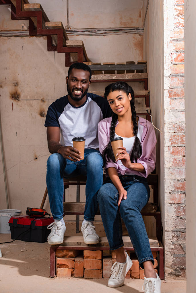 lachende paar zittend op een trap met papieren kopjes koffie tijdens renovatie van woning en camera kijken - Foto, afbeelding