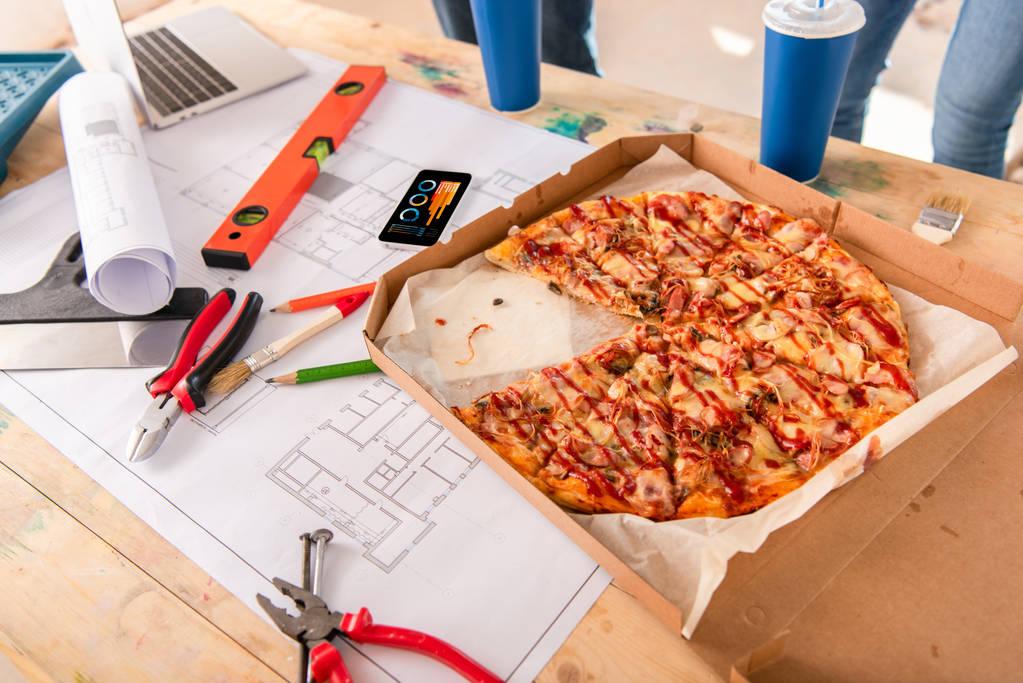 Nahaufnahme einer Schachtel mit Pizza, Werkzeug und Smartphone mit Diagrammen auf dem Bildschirm auf dem Bauplan - Foto, Bild