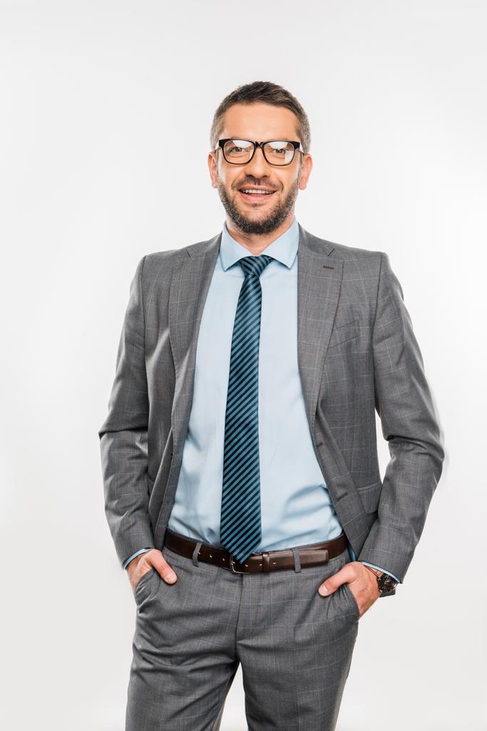handsome businessman in eyeglasses standing with hands in pockets and smiling at camera isolated on white  - Photo, Image