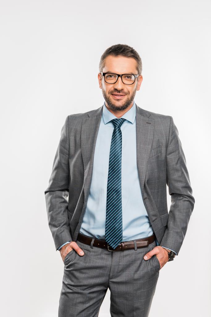 homem de negócios bonito em terno e óculos de pé com as mãos nos bolsos e sorrindo para a câmera isolada no branco
  - Foto, Imagem