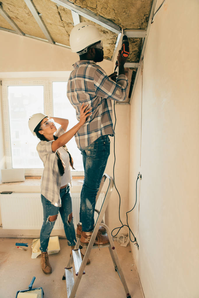 vista lateral do homem em chapéu duro e óculos de trabalho com broca de energia na escada, enquanto sua namorada de pé perto durante a renovação de casa
 - Foto, Imagem