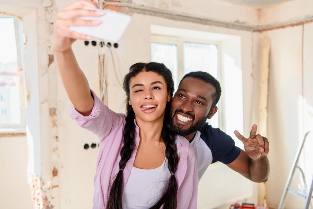 positiver afrikanischer Amerikaner zwinkert beim Selfie mit Freund, der Friedenszeichen auf Smartphone macht  - Foto, Bild