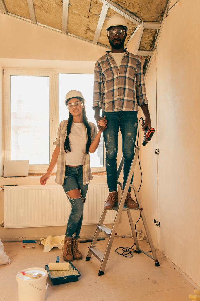 uomo africano americano in cappello duro e occhiali in piedi con trapano elettrico sulla scala mentre la sua ragazza in piedi vicino durante la ristrutturazione della casa
 - Foto, immagini