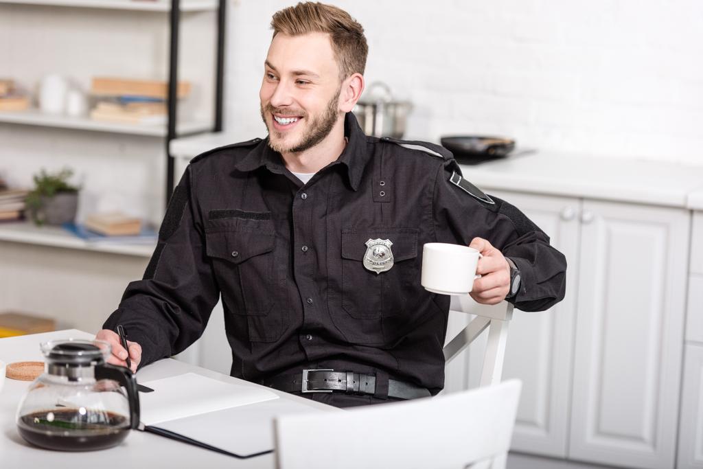 oficial de policía sonriente sentado en la mesa de la cocina, escribiendo en un cuaderno y tomando café
 - Foto, Imagen